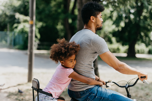 Carefree African American father and daughter cycling in the park. Mixed race family riding a bike together through the urban part of the city. A loving parent cycles around the block with his kid sitting in the children's seat.