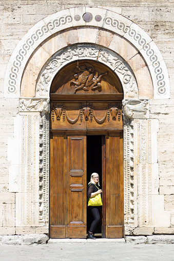 Bevagna, Umbria, Italy: Blond female tourist (active senior) exiting the Church of San Michele Arcangelo (close-up doorway). The 12th-century church is located on the main piazza, Piazza Silvestri, also known as Piazza Maggiore. Bevagna is part of Perugia Province.