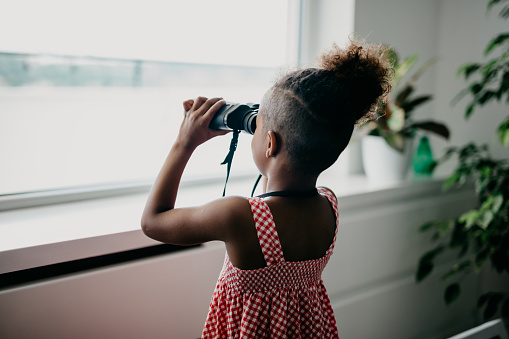 Adorable little african child wearing plaid dress is using binoculars to watch through the window.