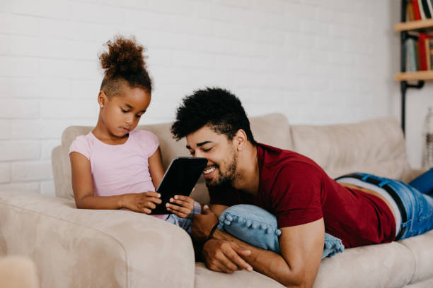 istock Single millennial parent is lying on  his belly on the sofa and watching his daughter playing video games on digital tablet. 814010216