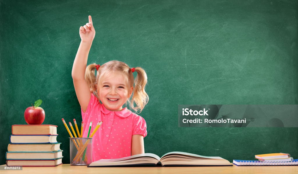 Happy Girl Raising Hand In The Classroom Little Student Sitting On The Desk With Blackboard and Books Child Stock Photo