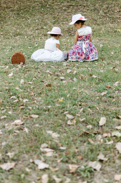two little sisters or friends sitting on the grass in nature, sisters on picnik, two little fashion model with beautiful dress and hat sitting outside, back view of two girls
