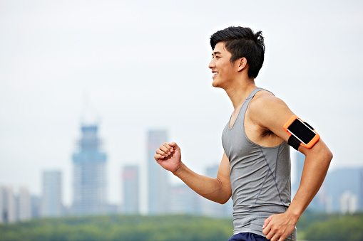 young asian male jogger with cellphone attached to arm running with skyline in the background.