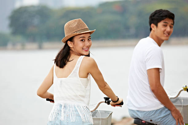 young asian couple turning to camera smiling while riding bicycle outdoors, slightly grainy