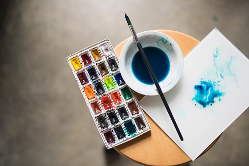 Artist's palette with painting colors, bowl with diluted blue paint and a paintbrush on the chair.