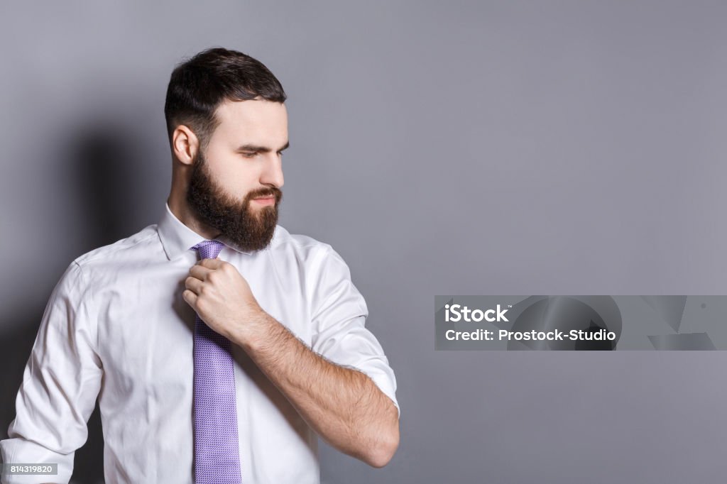 Handsome bearded businessman adjusting his tie Handsome bearded young businessman adjusting his tie with a serious thoughtful expression, looking away, gray studio background, copy space Adjusting Stock Photo