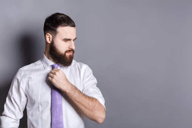 Handsome bearded businessman adjusting his tie Handsome bearded young businessman adjusting his tie with a serious thoughtful expression, looking away, gray studio background, copy space slenderman fictional character stock pictures, royalty-free photos & images