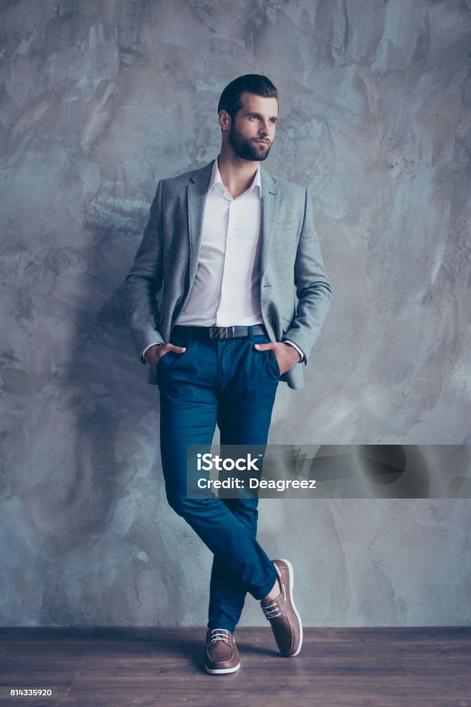 Full size portrait of stylish young bearded man standing on gray concrete background. He is in a suit, standing with crossed legs Men Stock Photo