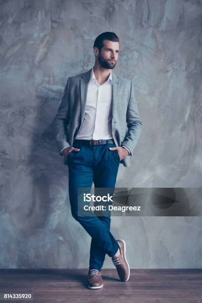 Full Size Portrait Of Stylish Young Bearded Man Standing On Gray Concrete Background He Is In A Suit Standing With Crossed Legs Stock Photo - Download Image Now