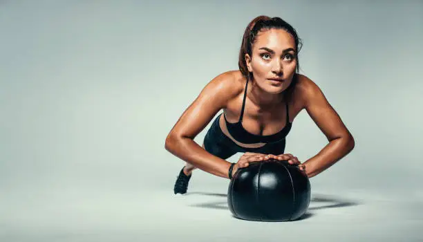 Horizontal shot of young fit woman doing push up on medicine ball. Fitness female exercising with a medicine ball on grey background.