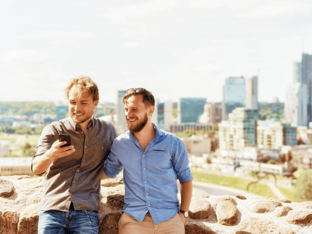 Smiling young caucasian buddies having fun while looking pictures at the mobile phone in front of city skyline as travel and friendship concept