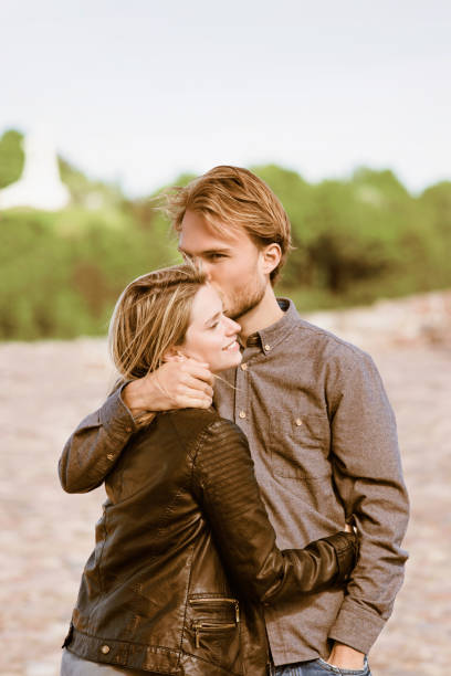 man embraces a woman in a park, covering her with his plaid shirt as the sun sets in the background
