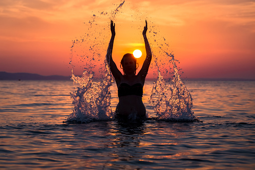 Beautiful female in ocean splashing water with hands. Sunset in the background. Nature and summer vacation concepts.