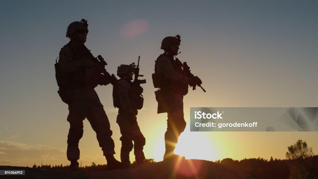 Squad of Three Fully Equipped and Armed Soldiers Standing on Hill in Desert Environment in Sunset Light. Armed Forces Stock Photo