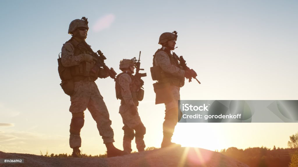 Squad of Three Fully Equipped and Armed Soldiers Standing on Hill in Desert Environment in Sunset Light. Iraq Stock Photo