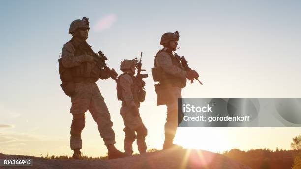 Squad Of Three Fully Equipped And Armed Soldiers Standing On Hill In Desert Environment In Sunset Light Stock Photo - Download Image Now