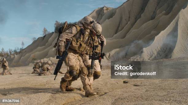 Soldier Carrying Injured One While Other Members Of Squad Covering Them During Military Operation In The Desert Stock Photo - Download Image Now