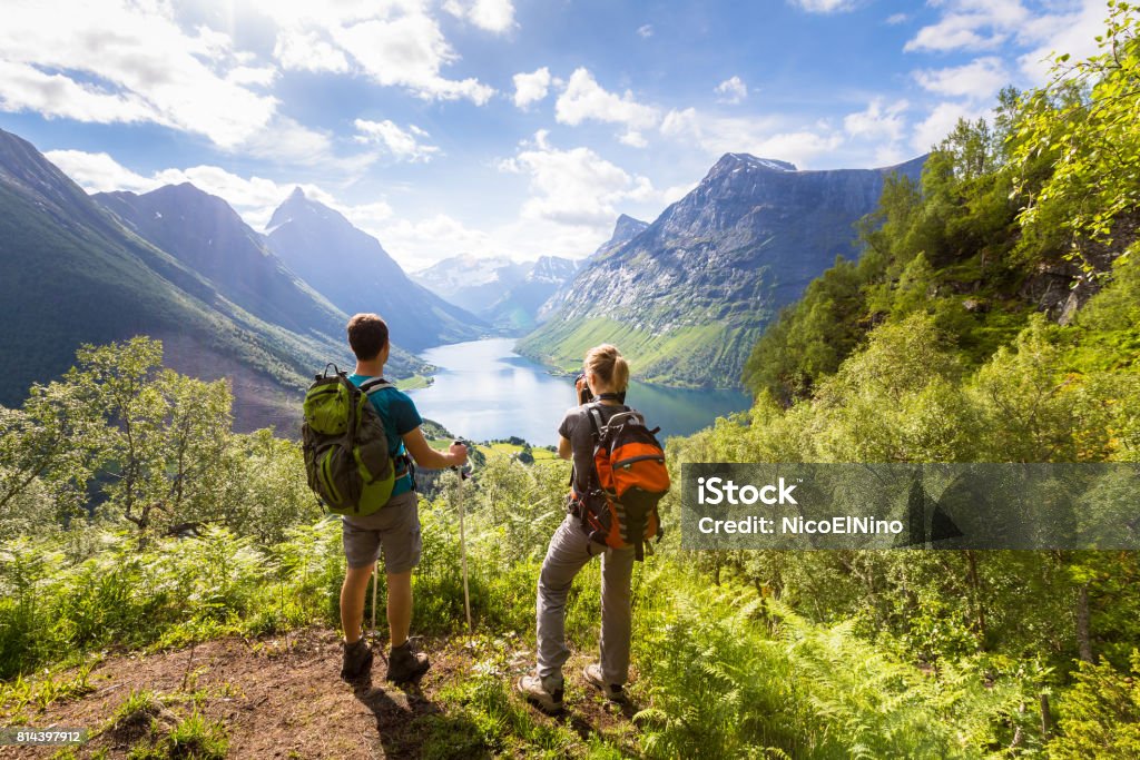 Two hikers at viewpoint  in mountains with lake, sunny summer Two hikers at viewpoint in the mountains enjoying beautiful view of the valley with a lake and sunny warm weather in summer, green trees around Hiking Stock Photo