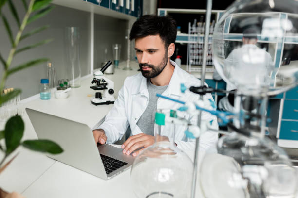 istock Focused young scientist in white coat using laptop in chemical lab 814397962