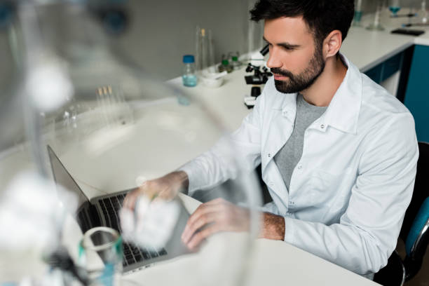 istock Serious bearded scientist using laptop in chemical laboratory 814398646