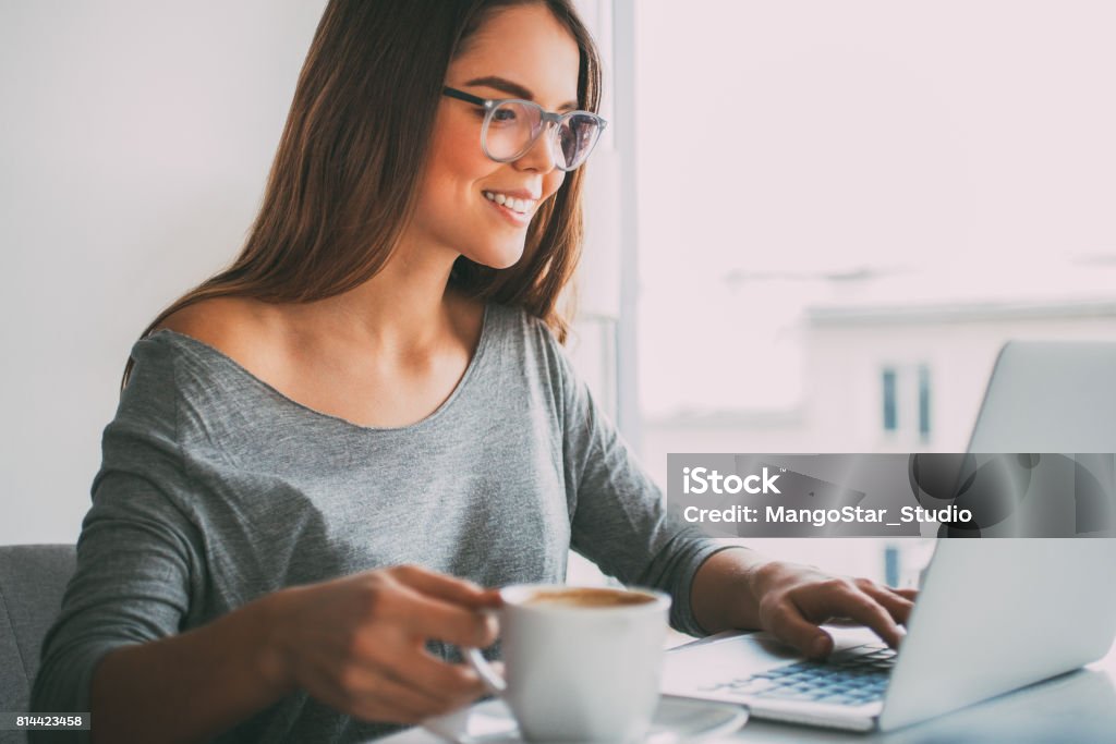 Happy businesswoman drinking coffee at laptop Portrait of happy casual young businesswoman wearing glasses sitting at cafe or office, drinking coffee while working on laptop and smiling Digital Tablet Stock Photo
