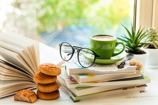 Coffee cup, open book, glasses, cookies and flower on window with bokeh. Reading and breakfast. Concept warm and cozy home interior