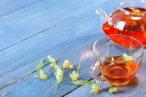 Limeflower tea in glass tea cup and teapot and lime flowers over blue wood table. Close-up