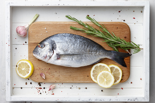Fresh dorado or gilthead bream cooking with lemon, garlic, herbs and spices on white wooden background
