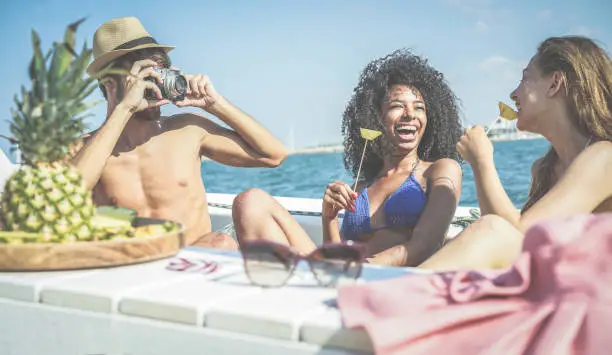 Photo of Happy friends having fun and eating fresh pineapple fruit at boat party - Young people taking photos in caribbean sea tour - Youth, tropical, travel and summer vacation concept - Focus on left guys