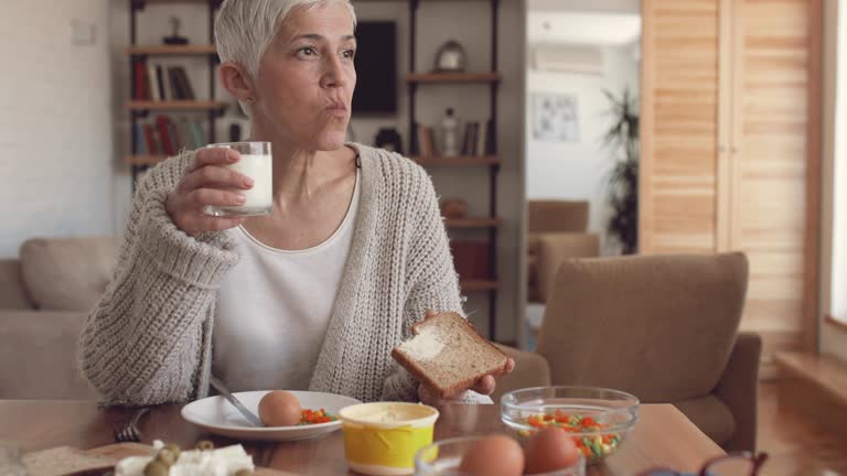 Mature woman eating a breakfast at dining table at home.