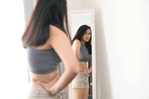 Body image issues A young woman looking at her reflection in the mirror, unhappy with what she is seeing. eating disorder stock pictures, royalty-free photos & images