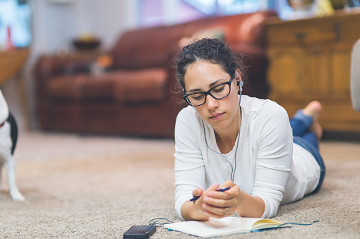 Young ethnic woman in living room relaxes and listens to exciting podcast on her phone while she writes in her journal