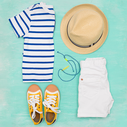 Child's striped t-shirt, denim shorts, accessories, yellow shoes and straw hat on turquoise wooden background. Top view. Flat lay. Kid's summer clothes collage