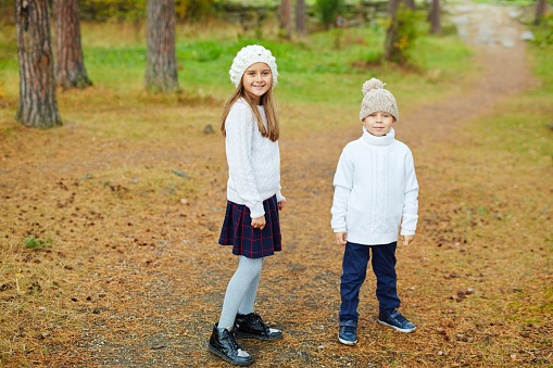 Brother and sister posing to camera, smiling outdoors  in pine forest enjoying the walk on warm autumn day , both dressed in similar knit clothes