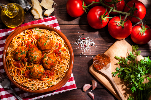 Top view of a plate filled with spaghetti and meatballs with some ingredients for preparation like tomatoes, parsley, garlic, olive oil and Parmesan cheese shot on rustic wooden table. Low key DSRL studio photo taken with Canon EOS 5D Mk II and Canon EF 100mm f/2.8L Macro IS USM