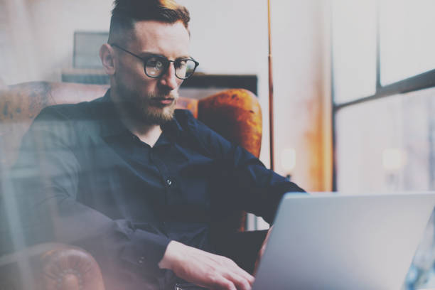 istock Bearded young businessman in eyeglasses working in office.Man using contemporary notebook while sitting in vintage chair.Panoramic windows on the blurred background. Horizontal,flare effect. 815160134