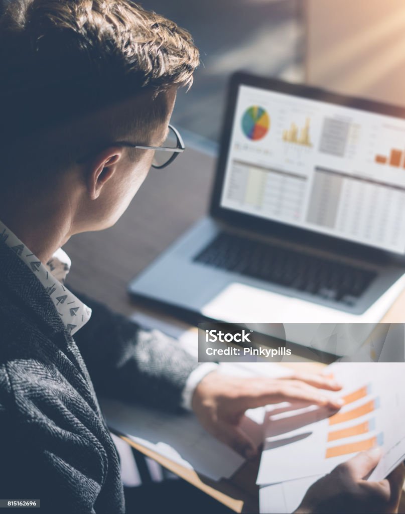 Young finance market analyst in eyeglasses working at sunny office on laptop while sitting at wooden table.Businessman analyze document in his hands.Graphs and diagramm on notebook screen.Vertical. Young finance market analyst in eyeglasses working at sunny office on laptop while sitting at wooden table.Businessman analyze document in his hands.Graphs and diagramm on notebook screen.Vertical Finance Stock Photo