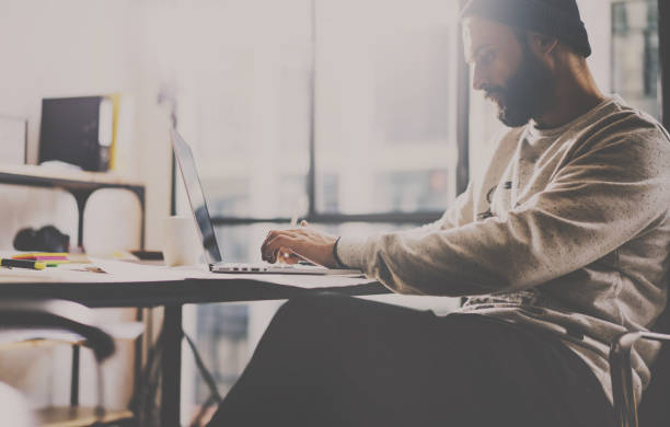 istock Bearded designer typing on laptop keyboard at modern coworking studio at the wooden table.Panoramic windows on blurred background.Horizontal. 815165784