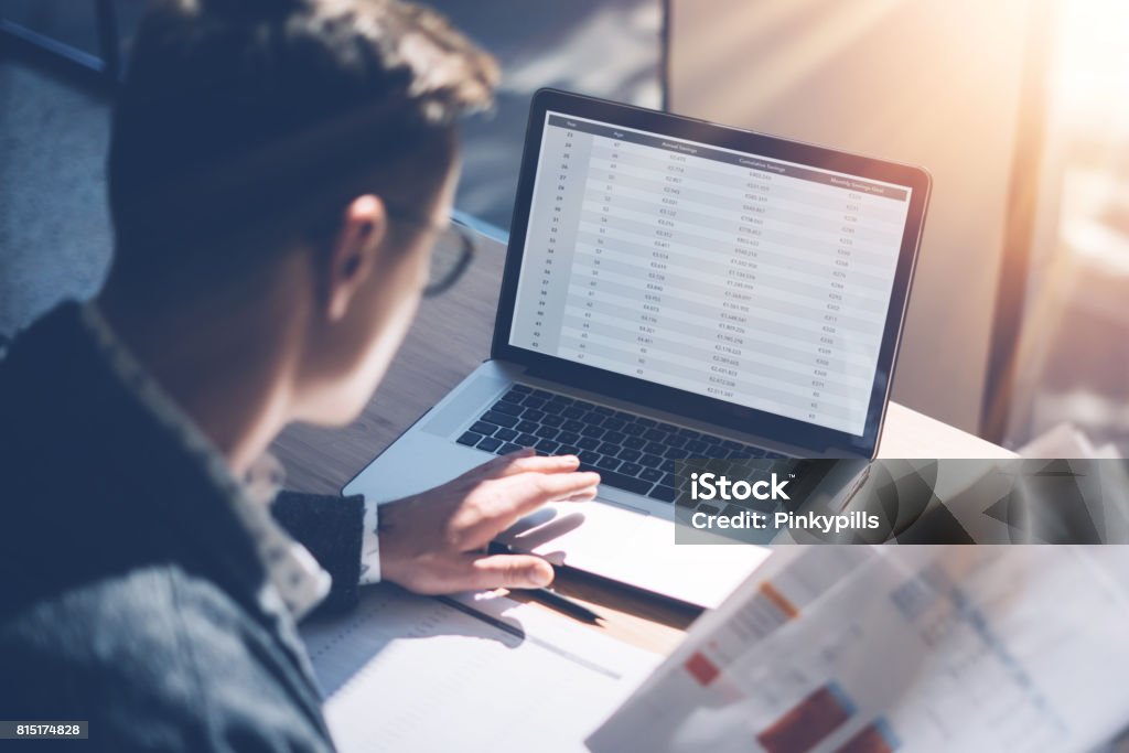 Closeup view of banking finance analyst in eyeglasses working at sunny office on laptop while sitting at wooden table.Businessman analyze stock report on notebook screen.Blurred background,horizontal. Closeup view of banking finance analyst in eyeglasses working at sunny office on laptop while sitting at wooden table.Businessman analyze stock report on notebook screen.Blurred background,horizontal Bank - Financial Building Stock Photo