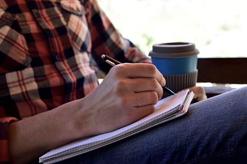 closeup of a young caucasian man wearing jeans and a plaid shirt writing with a pen in a notebook in the porch of a house or a ranch