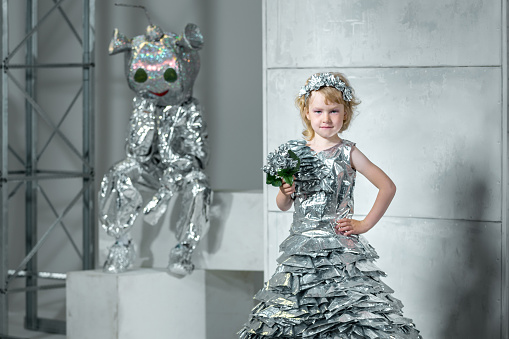 Two children (beautiful preschool girl and elementary age boy) are showing the clothes from secondary raw and recycling materials. The girl dressed in the princess dress made from silver foil is standing with her hand at the waist. She is smiling looking at the camera. The boy dressed in the the robot's helmet and the robot costume is sitting on a the pedestal stand behind the girl. Studio shooting on white grunge wall background
