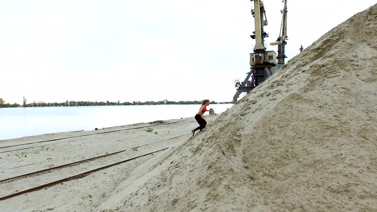 Young woman athlete climbs a sandy mountain. On the background can be seen the river, cargo cranes, cargo port, dawn