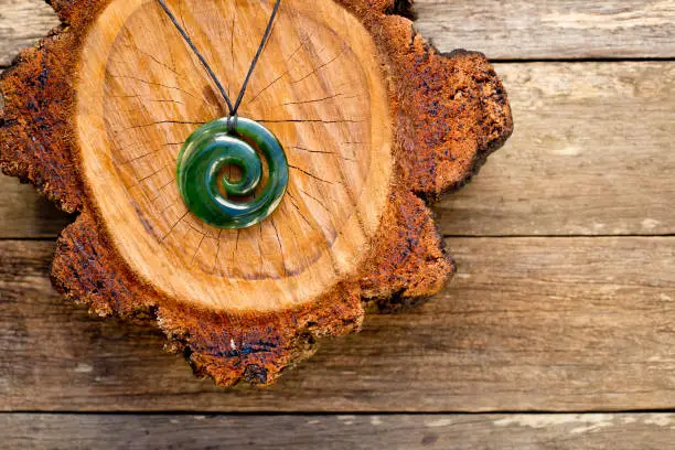 Maori New Zealand Greenstone pounamu jade Koru (spiral fern) shape pendant on cross section of timber log on wooden plank background