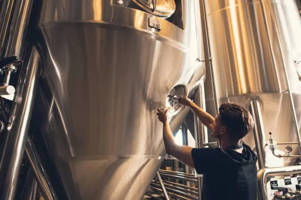 Young man working in beer manufacturing factory. Brewer working with industrial equipment at the brewery.