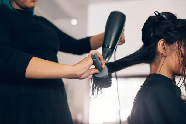 Female hairdresser using blower and brush to dry hair. Hair stylist using dryer on woman wet hair in salon.