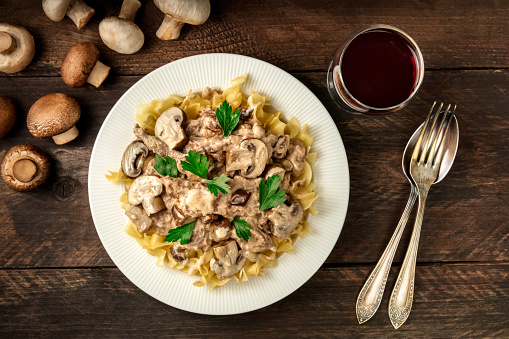 Mushroom beef stroganoff, served with the typical egg pasta, with cremini and champignons, with a glass of red wine, a fork, and a spoon, shot from above on a dark rustic texture with a place for text