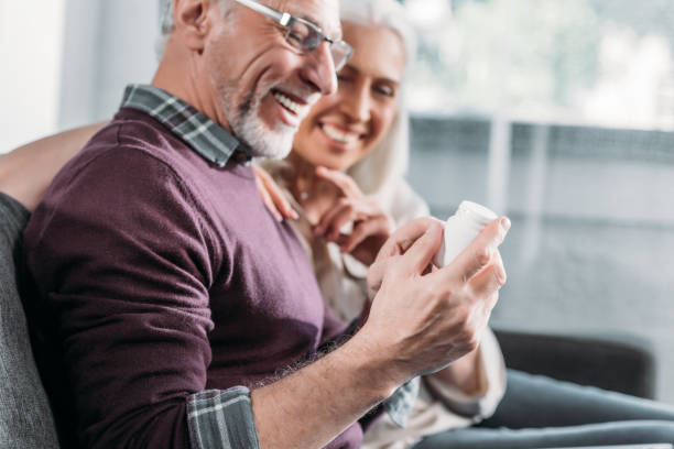 sorrindo casal sênior com frasco de comprimidos, sentado no sofá em casa - vitamin pill nutritional supplement pill bottle - fotografias e filmes do acervo