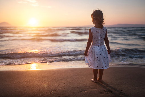 Adorable girl watching sunset standing at sandy beach near sea