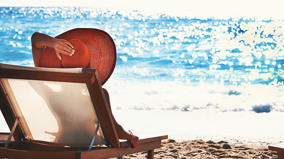 Closeup rear view of a woman relaxing on a deck chair on a beach. It's sunny summer day and she's enjoying the peacefulness and the sound of the waves crushing on the shore. She's wearing big red hat.
