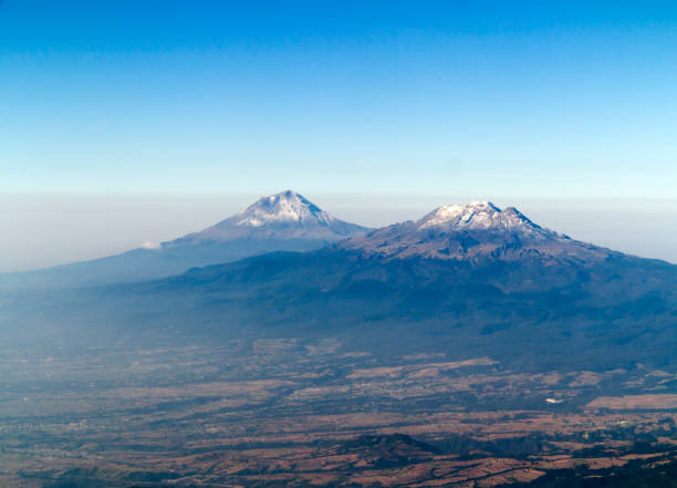 Volcanos Popcatepetl and Iztaccihuatl, Mexico Active Popocatepetl volcano (5.452 meters) with a bit of smoke rising above the smog and clouds of Mexico City in the Valley of Mexico. "Popo" is Mexico's second highest mountain. To it's right is Iztaccihuatl, an extinct volcano. They are a mere 70 km (44 mi) to the southeast of Mexico City and is often visible from the capital, depending on atmospheric conditions. Mexico. popocatepetl volcano photos stock pictures, royalty-free photos & images